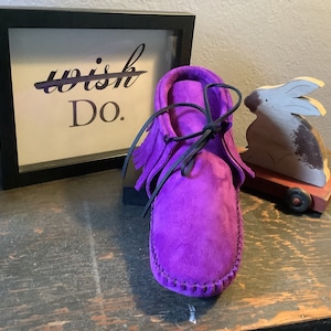 May include: Purple suede moccasins with black laces and fringe. The moccasins are sitting on a wooden surface with a framed sign that says "Do."