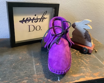 May include: Purple suede moccasins with black laces and fringe. The moccasins are sitting on a wooden surface with a framed sign that says "Do."