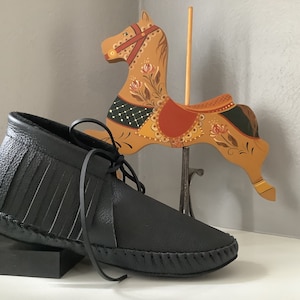 May include: A pair of black leather moccasins with fringe detailing. The moccasins are tied with a black leather lace and have a stitched seam around the sole.