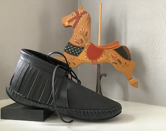 May include: A pair of black leather moccasins with fringe detailing. The moccasins are tied with a black leather lace and have a stitched seam around the sole.
