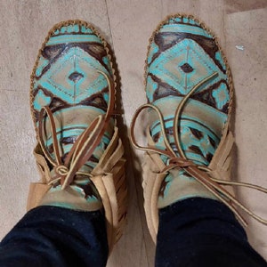 May include: A pair of brown leather moccasins with turquoise and brown geometric patterns. The moccasins have laces that tie at the ankle.