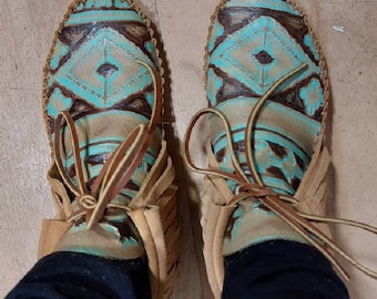May include: A pair of brown leather moccasins with turquoise and brown geometric patterns. The moccasins have laces that tie at the ankle.