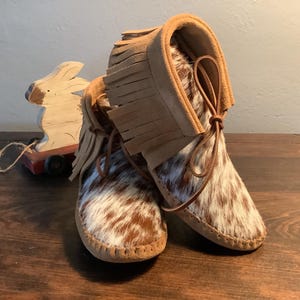 Peut inclure: Une paire de mocassins en cuir de vachette marron et blanc avec des détails en franges. Les mocassins ont une bride en cuir beige et sont attachés avec un lacet en cuir marron.
