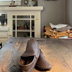 May include: A pair of dark brown leather moccasins featuring a textured grain finish, a rounded toe, and visible whipstitch detailing along the outer edges of the sole and the upper vamp, presented in a slip-on style with a low-profile construction.