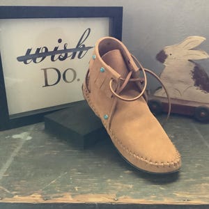 May include: Tan leather moccasin boots with turquoise accents and a fringe detail. The boots are tied with a brown leather lace. The boots are sitting on a wooden surface.