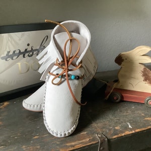 May include: White leather moccasins with brown leather laces and turquoise and black beads. The moccasins have fringe on the sides and are tied at the ankle.