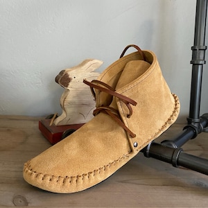 Pu&ograve; includere: Mocassini in camoscio beige con lacci marroni e una cucitura decorativa attorno alla suola. I mocassini sono su una superficie di legno.