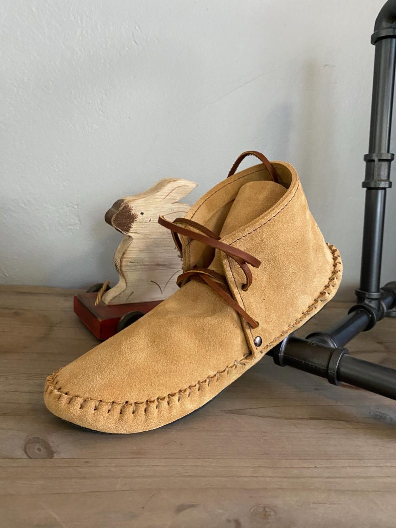 Pu&ograve; includere: Mocassini in camoscio beige con lacci marroni e una cucitura decorativa attorno alla suola. I mocassini sono su una superficie di legno.