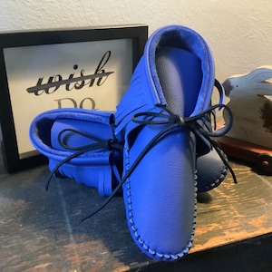 May include: A pair of blue leather moccasins with black laces tied in a bow. The moccasins have fringe detailing and are sitting on a wooden surface.