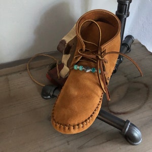 May include: A tan suede moccasin boot with leather laces and turquoise beads. The boot has a stitched sole and a high ankle. A decorative item is behind the boot.