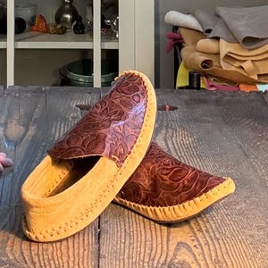 May include: A pair of slip-on moccasins featuring a dark brown leather upper with an embossed floral pattern and a light tan suede sole. The shoes are constructed with visible whipstitching along the edges, connecting the textured leather top to the soft, light-colored suede base.