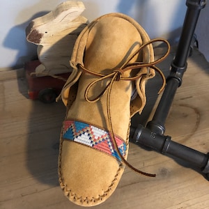 May include: Tan leather moccasins with a colorful beaded band around the ankle. The moccasins are tied with a brown leather lace.