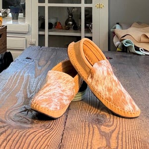 May include: A pair of slip-on moccasins featuring a tan suede base and a textured, hair-on-hide upper with a mottled white and brown pattern. The shoes have visible whipstitch detailing along the edges and a rounded toe construction.