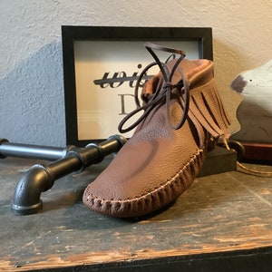 May include: A brown leather moccasin with fringe and a lace-up closure. The moccasin is sitting on a wooden surface.