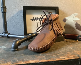 May include: A brown leather moccasin with fringe and a lace-up closure. The moccasin is sitting on a wooden surface.