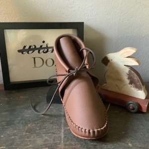 May include: Brown leather moccasin style ankle boot with laces tied in a bow. The boot is sitting on a wooden surface with a framed print that says "Do" and a wooden toy rabbit on a red wagon.