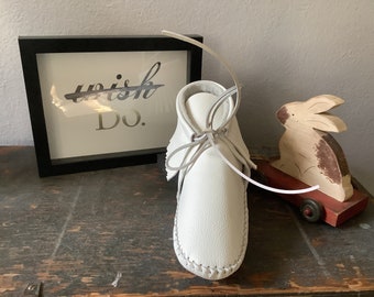 May include: White leather moccasin style baby shoes with laces tied in a bow. The shoes are on a wooden surface with a framed sign that says "wish Do."