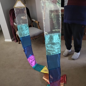 May include: A long, vertical hanging decoration featuring two parallel, transparent plastic panels connected by a horizontal wooden rod. Each panel displays a series of rectangular, metallic-finish fabric sections in shades of teal, blue, purple, and gold, topped with a white triangular header trimmed in gold.