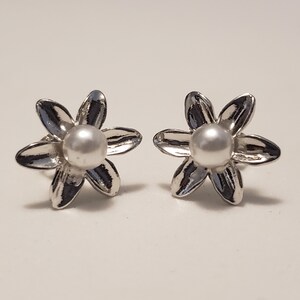 May include: A pair of silver-toned flower stud earrings. Each earring features six petals surrounding a white pearl center. The earrings are set against a white background, highlighting their reflective, metallic finish.
