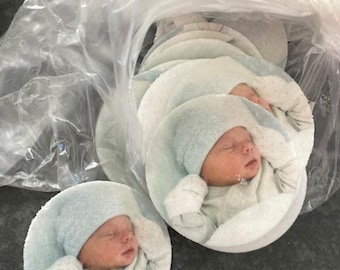 May include: A set of round, white coasters with a baby's face printed on each one. The baby is wearing a light blue hat and a white onesie. The coasters are packaged in a clear plastic bag.