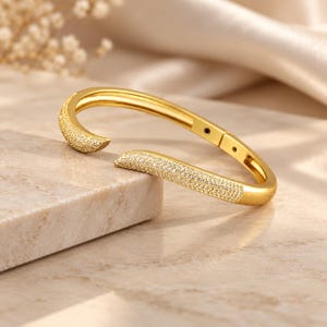 May include: A gold-toned open cuff bracelet featuring a curved, bypass-style design. The two tapered ends are encrusted with numerous small, clear pavé-set stones that catch the light, while the inner band remains smooth and polished gold.