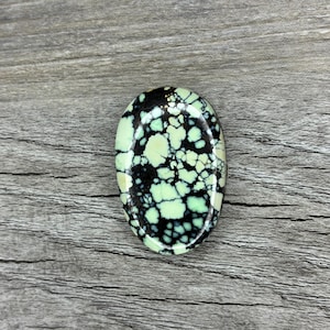 May include: An oval-shaped gemstone with a polished surface. The stone features a striking pattern of black and light green, with the green appearing in irregular, cloud-like shapes. The gemstone is set against a wooden background.