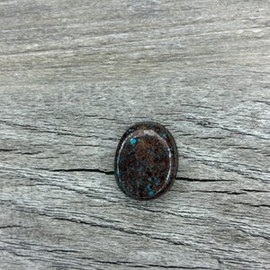 May include: Oval-shaped gemstone with a dark brown base and speckled turquoise blue inclusions. The polished stone has a smooth surface and is set against a gray wooden background. The gemstone is a natural stone.