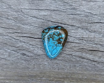 Può includere: Un cabochon turchese con matrice marrone, tagliato a forma di lacrima. La pietra ha una superficie liscia e lucida ed è posizionata su uno sfondo di legno.