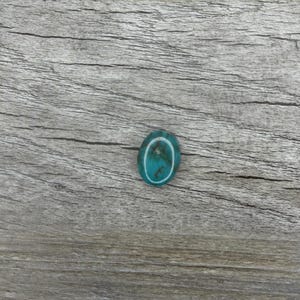 May include: Oval-shaped turquoise gemstone with a white border. The polished stone displays a vibrant blue-green color with natural veining. The gemstone is set against a weathered, gray wooden surface.
