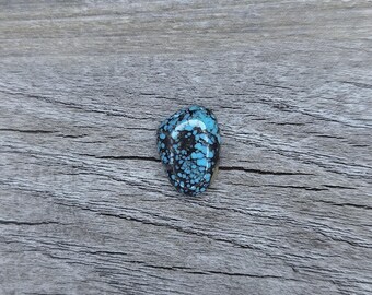 May include: A blue and black turquoise cabochon with a spiderweb pattern. The stone is set on a grey wooden surface.