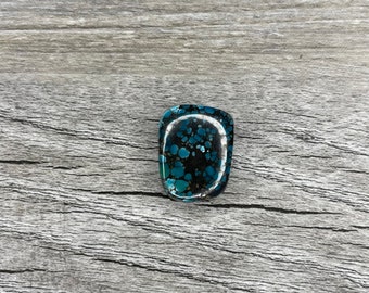 May include: A turquoise cabochon with black and white markings. The stone is set in a silver bezel and has a smooth, polished surface.