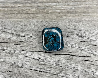May include: A square-shaped turquoise cabochon with a black matrix and a blue and white pattern. The stone is set in a clear resin.