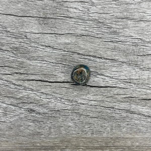 Pode incluir: Um pequeno botão redondo e decorativo com uma superfície texturizada. O botão apresenta uma mistura de cores azul, castanho e bege. O botão está sobre um fundo de madeira cinzenta.