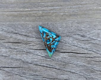 May include: A triangular turquoise cabochon with a brown matrix. The turquoise is a vibrant blue with brown and black veins. The cabochon is set on a wooden surface.