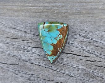 May include: A triangular turquoise gemstone with a polished surface. The stone features a vibrant blue color with brown matrix patterns. The gemstone is set against a gray wooden background, highlighting its natural beauty. This gemstone is ideal for jewelry making.