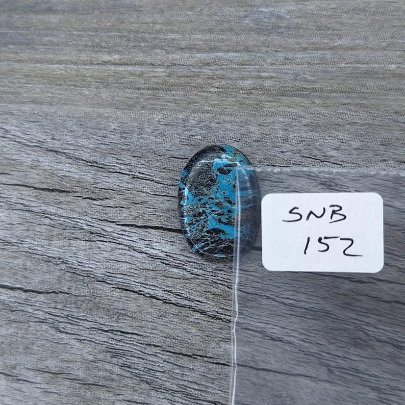 May include: A close-up of a blue and black oval cabochon gemstone with a white label that says "SNB 1572".