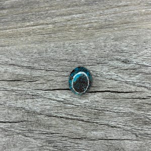 May include: Oval-shaped turquoise gemstone with a dark brown center and a white border. The gemstone is set against a weathered, gray wooden surface. The stone's vibrant blue and brown colors create a striking contrast.