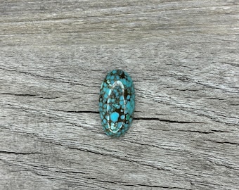 May include: An oval turquoise gemstone, displaying a mottled pattern of blue, brown, and white. The polished stone is set against a weathered, grey wooden surface. This gemstone is suitable for jewellery making and crafting.