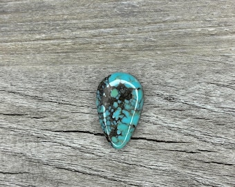 Può includere: Una gemma turchese lucidata a forma di goccia. La pietra presenta un sorprendente motivo di blu chiaro e marrone scuro. È posta su uno sfondo di legno grigio, che ne mette in risalto i colori e le texture naturali. La gemma è lunga circa 3 cm.