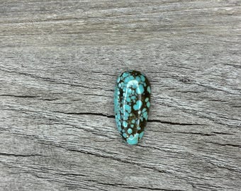 May include: A polished, teardrop-shaped gemstone with a turquoise and brown mottled pattern. The stone is set against a weathered, gray wooden surface, highlighting its natural colours and textures. The gemstone is approximately 2.5 cm in length.