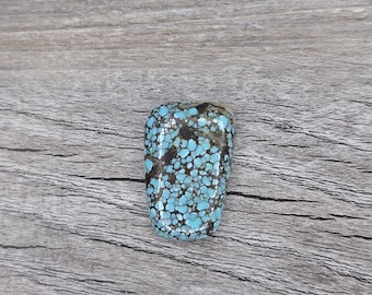 May include: A polished, rectangular turquoise gemstone with a speckled pattern of light blue and dark brown spots. The stone is set against a grey wooden background, showcasing its natural beauty. This gemstone is ideal for jewelry making.