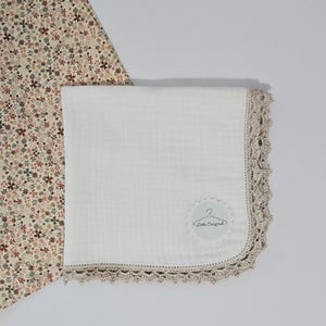 May include: A white baby blanket with a crocheted beige border. The blanket has a soft, textured appearance and features a small, circular tag with the text "Little Original". A floral patterned fabric is visible in the background.