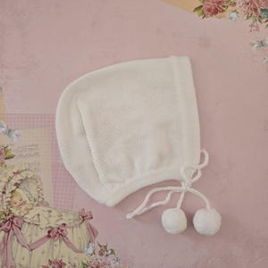 Puede incluir: Un gorro de bebé blanco tejido, con una corona redondeada y un borde cuadrado. El gorro tiene dos pompones blancos sujetos con una cuerda. El gorro está hecho de un material suave y texturizado.