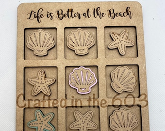 May include: Wooden tic-tac-toe game with a beach theme. The game board features squares with seashell and starfish designs. The text "Life is Better at the Beach" and "Crafted in the 603" are engraved on the board. Includes a separate starfish game piece.