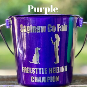 May include: A metallic purple bucket with a silver handle. The front features the text "Saginaw Co Fair" at the top and "FREESTYLE HEELING CHAMPION" at the bottom, with a silhouette graphic of a person standing next to a sitting dog in between.