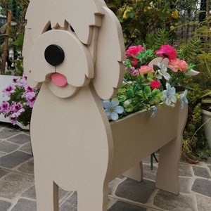 May include: A beige wooden dog-shaped planter filled with colorful artificial flowers. The dog has a black nose, pink tongue, and a shaggy head. The planter is on a stone patio.
