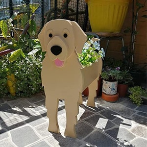 May include: A light tan wooden dog planter with a black nose and pink tongue. The dog planter is standing and has a container on its back filled with green plants and blue flowers. The planter is outdoors.