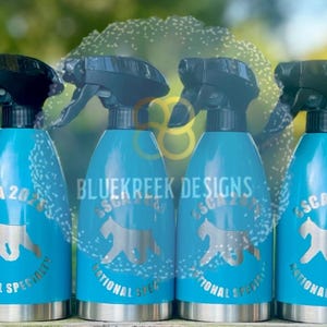 K&ouml;nnte beinhalten: Eine Reihe von zehn leuchtend blauen Spr&uuml;hflaschen mit schwarzen Spr&uuml;hk&ouml;pfen. Jede Flasche zeigt eine silberne Grafik einer Hundesilhouette und den Text "ASCA 2024 NATIONAL SPECIALTY". Der Markenname "BLUEKREEK DESIGNS" ist deutlich zu erkennen.
