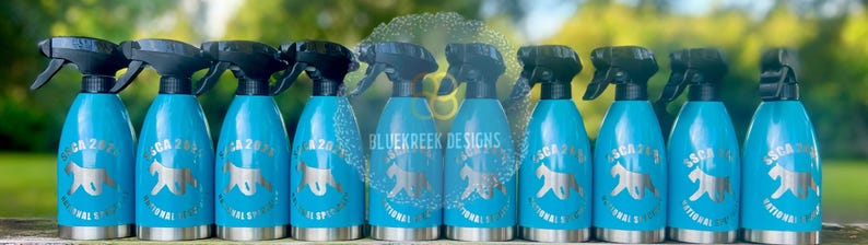 K&ouml;nnte beinhalten: Eine Reihe von zehn leuchtend blauen Spr&uuml;hflaschen mit schwarzen Spr&uuml;hk&ouml;pfen. Jede Flasche zeigt eine silberne Grafik einer Hundesilhouette und den Text "ASCA 2024 NATIONAL SPECIALTY". Der Markenname "BLUEKREEK DESIGNS" ist deutlich zu erkennen.