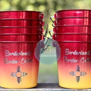 May include: Two stacks of red and yellow ombre metal buckets with silver wire handles. Each bucket features the text Borderlands Border Collies and a Zia sun symbol in silver. The finish is glittery, transitioning from deep red at the top to yellow.